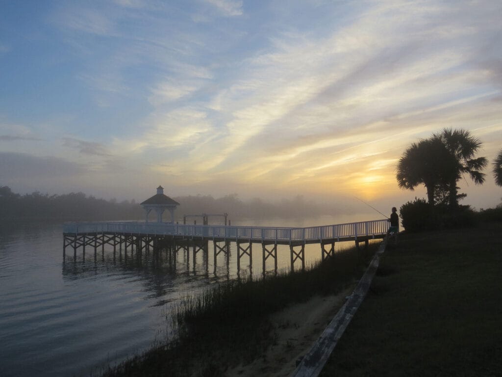 Peaceful lakeside sunrise with wooden dock, gazebo, palm tree, and silhouette by calm reflective water.