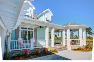 Light blue coastal house with white trim, gabled roof, and wraparound porch under sunny sky.