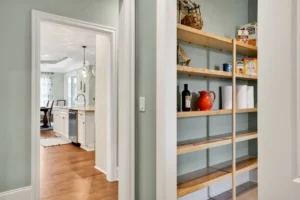 Modern open-concept kitchen with light wood pantry shelves, sage-green walls, and natural sunlight.