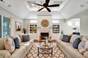 Bright elegant living room with white fireplace, tray ceiling, beige sofas, and built-in shelves.
