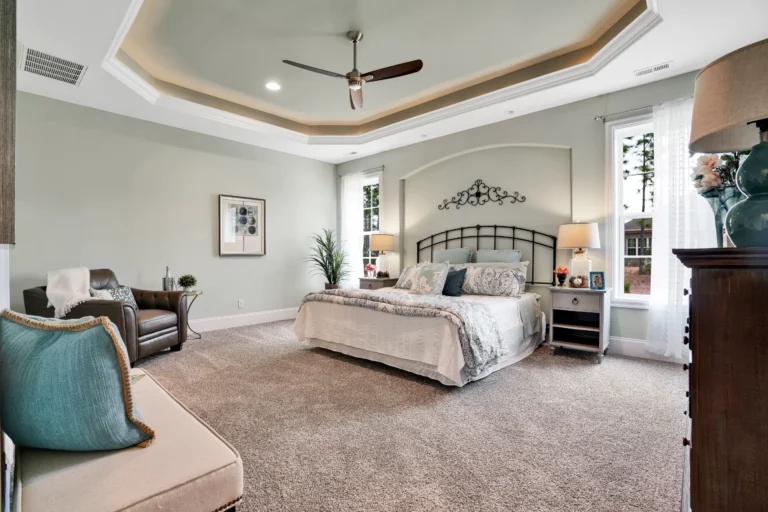 Elegant master bedroom with tray ceiling, neutral colors, cozy seating, and soft natural lighting.
