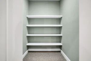 Modern minimalist storage nook with white floating shelves, soft green walls, and beige carpet flooring.