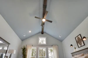 Bright modern farmhouse bedroom with vaulted ceiling, wood beam, ceiling fan, and large sunlit windows.