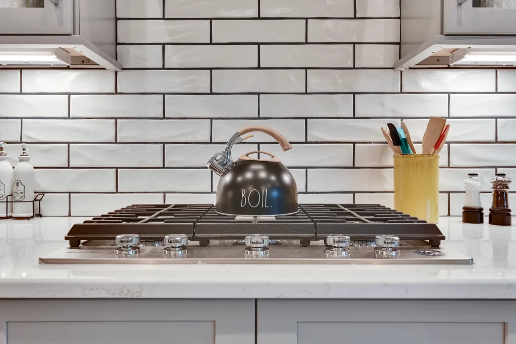 Modern white kitchen with stainless steel gas cooktop, black kettle, and yellow utensil holder.