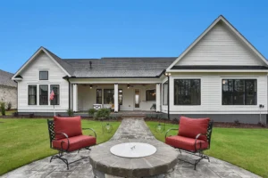 Modern farmhouse backyard with white exterior, stone fire pit, red-cushioned chairs, and lush lawn.