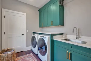 Modern teal laundry room with white countertops, brass accents, and front-loading washer and dryer.