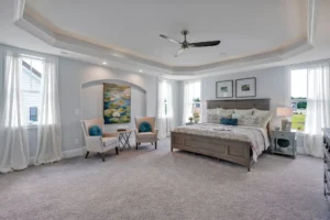 Modern gray primary bedroom with tray ceiling, wooden bed, cozy sitting area, and natural light.