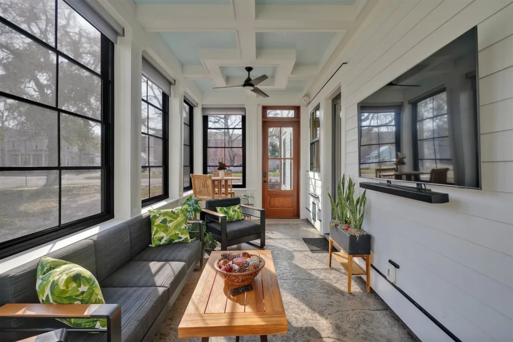 Bright modern sunroom with gray sofa, wood table, black windows, and green plant accents.