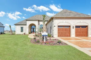 Modern stone house with wooden garage doors, manicured lawn, and bright blue sky.