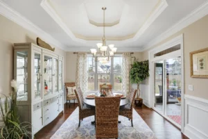 Elegant dining room with natural light, woven chairs, wood table, and chandelier under tray ceiling.
