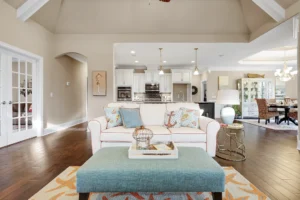 Bright coastal living room with white sofa, teal ottoman, open kitchen, and airy beach-inspired design.