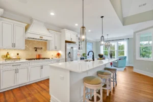 Modern white kitchen with quartz island, hardwood floors, pendant lights, and stainless steel appliances.