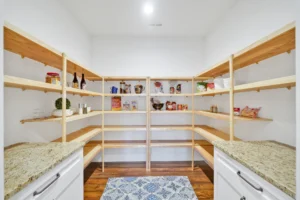 Bright modern pantry with light wood shelves, white walls, granite countertops, and blue patterned rug.