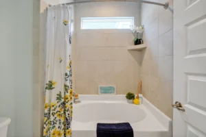 Modern beige bathroom with white tub, yellow floral shower curtain, blue towel, and natural light.