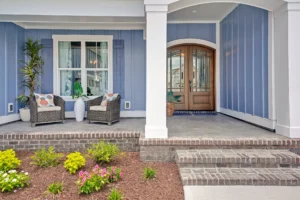 Coastal blue front porch with wooden double doors, white columns, wicker chairs, and blooming flowers.