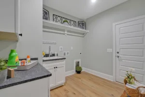 Modern light gray laundry room with white cabinets, dark countertop, wood floors, and decorative greenery.