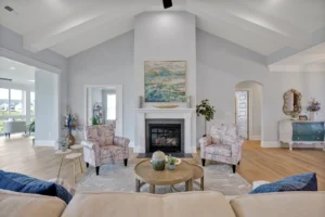 Bright coastal living room with vaulted ceiling, white fireplace, patterned chairs, and natural light.