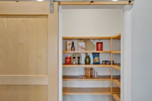 Modern wooden pantry with sliding barn door and neatly organized shelves in a bright kitchen.