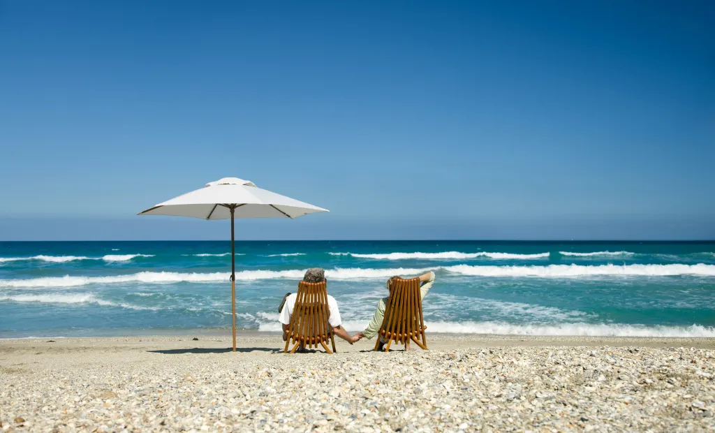 The New Coastal Haven: Choosing NC Over Florida 2 A couple sitting in wooden chairs under an umbrella on a sandy beach looking at the ocean.