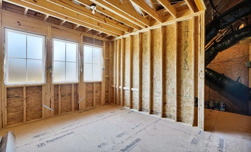 Rafters vs. Roof Trusses: Why Liberty Homes Chooses Site-Built Roof Framing 1 Unfinished attic room under construction with exposed wood framing, plywood walls, subflooring, and three frosted windows.